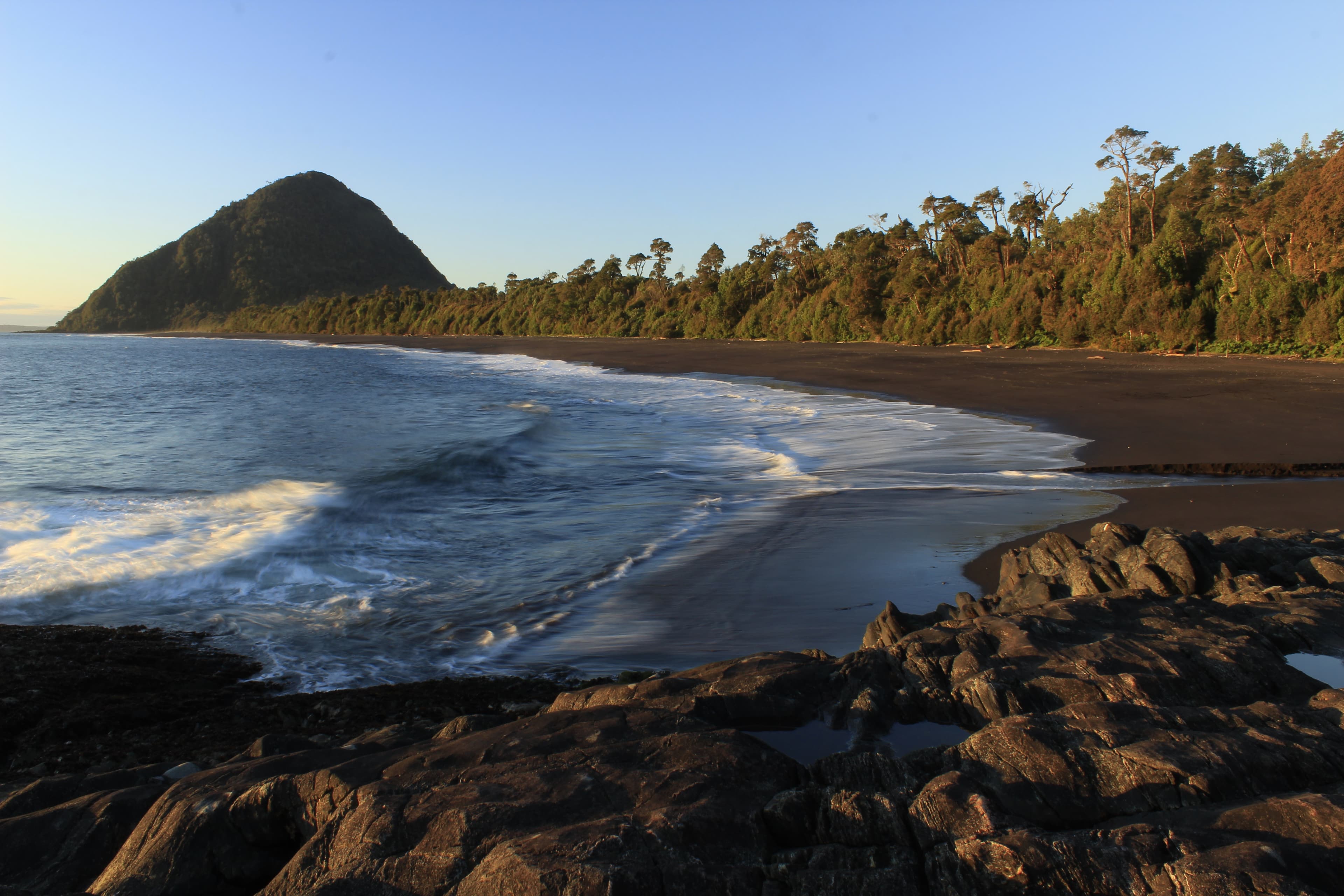 Playa Santa Bárbara al atardecer, con arena oscura, rocas en primer plano y un cerro boscoso cerrando la bahía